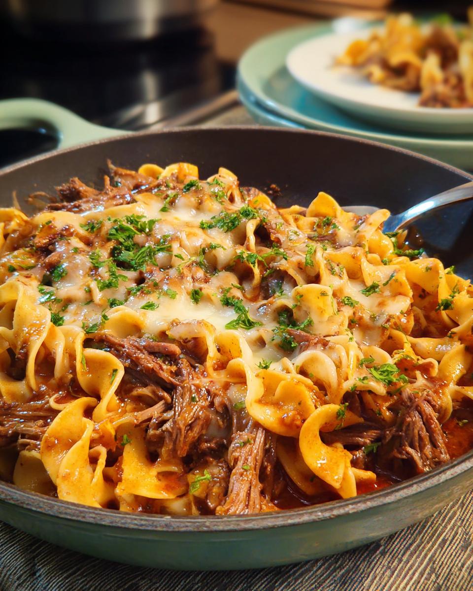 A close-up of a skillet filled with tender shredded beef, wide egg noodles, and melted cheese, garnished with fresh parsley. A classic dinner recipe.