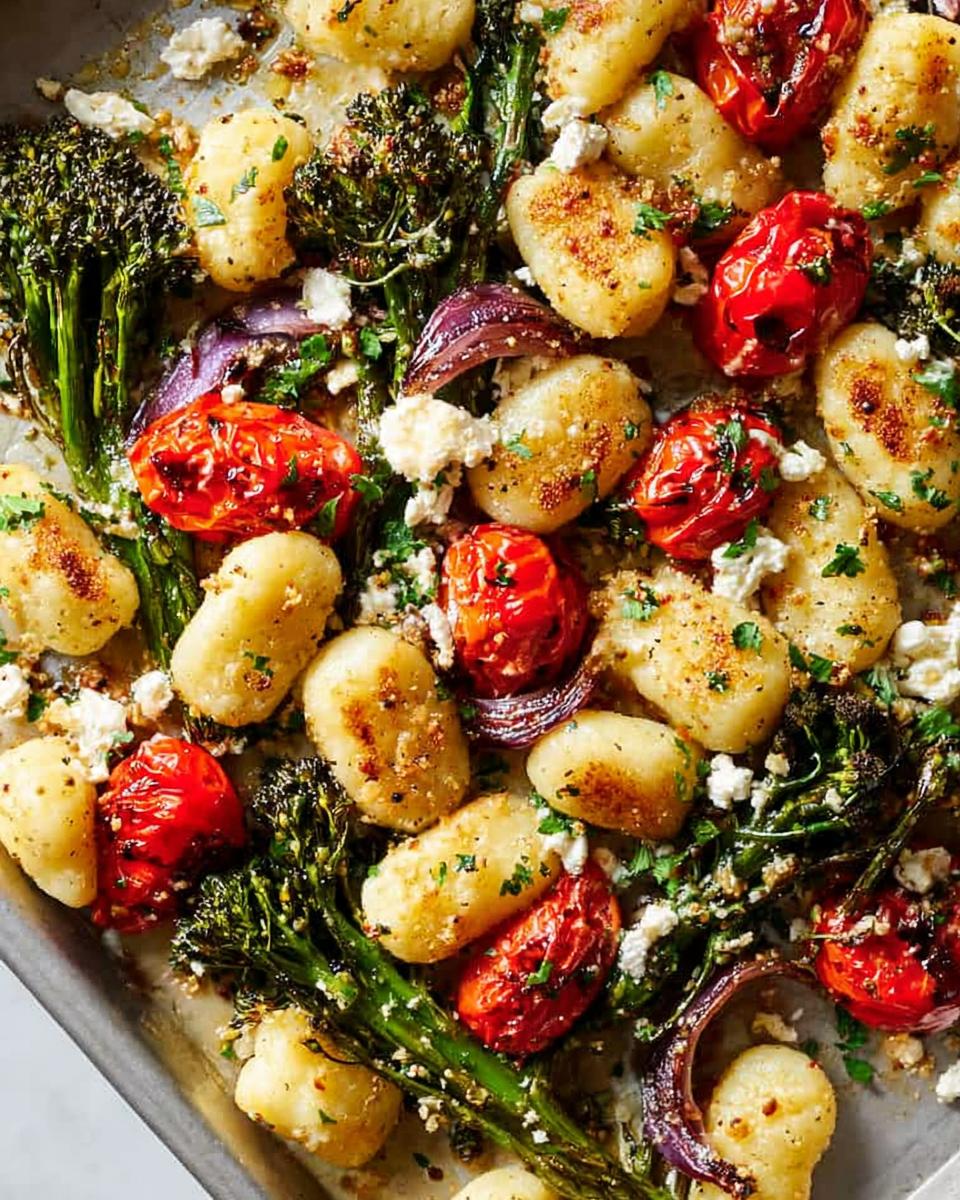 A close-up of a baking sheet filled with golden-brown gnocchi, roasted cherry tomatoes, broccolini, and red onion, topped with crumbled feta and herbs.