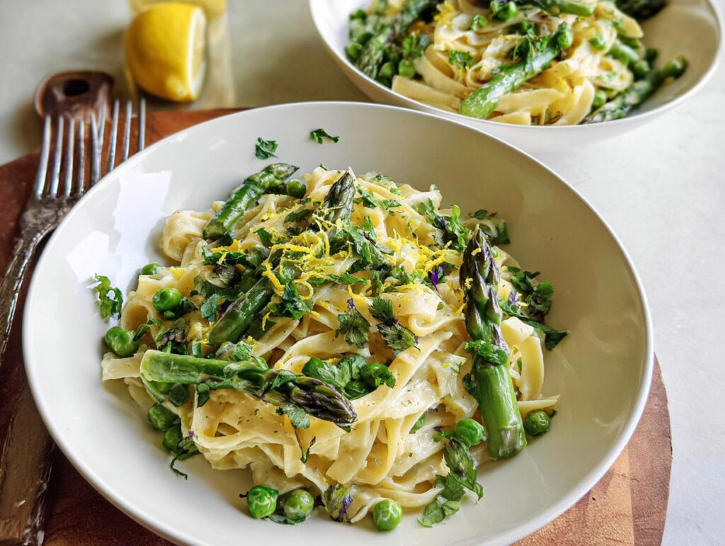 A bowl of creamy fettuccine pasta with fresh asparagus and peas, topped with lemon zest and herbs. A simple dinner recipe at home.